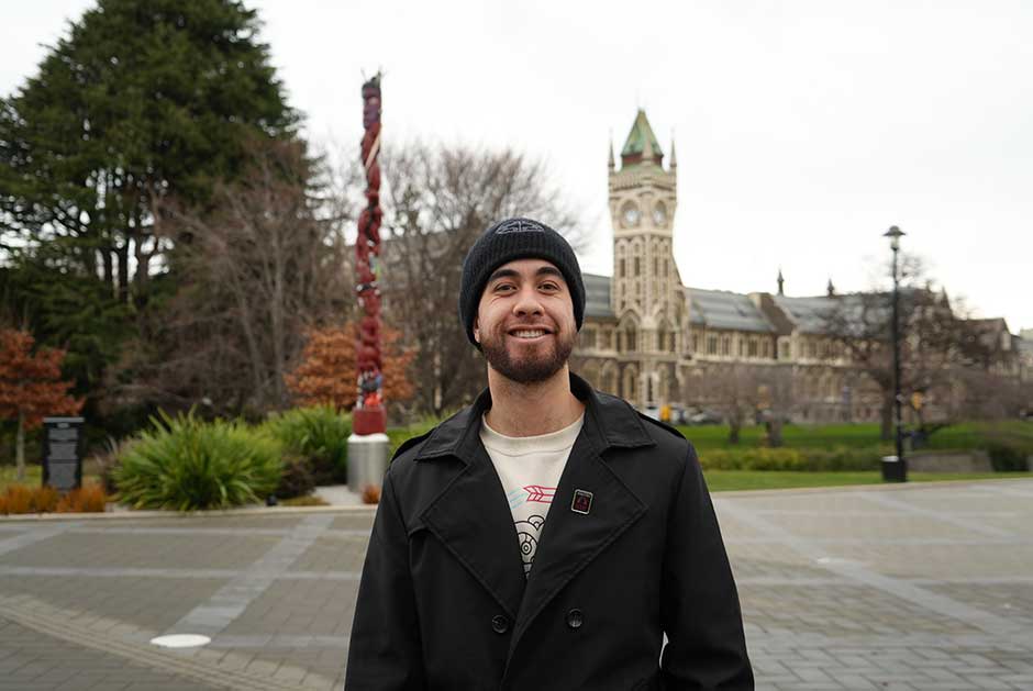 a young man standing in front of the Pou and Clocktower, smiling on an overcast day