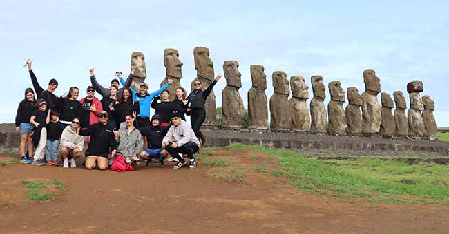 a group of people, crowded together, smiling and some waving at the camera, with a row of large stone monuments behind them