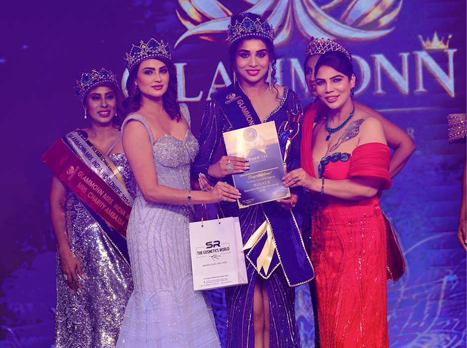 four women standing on stage, three wearing tiaras, one gifting another a certificate