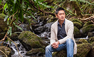 Joon Kim sitting on a rock in a forest next to a stream