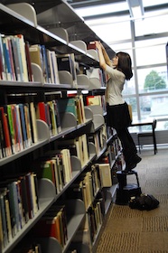 Photo of a young woman standing on a step stool arranging books on a high bookshelf 