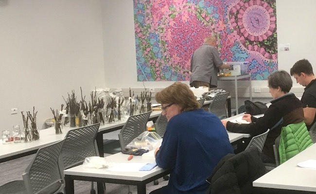 Room of people mostly sitting at desks with jars filled with sticks at the front.