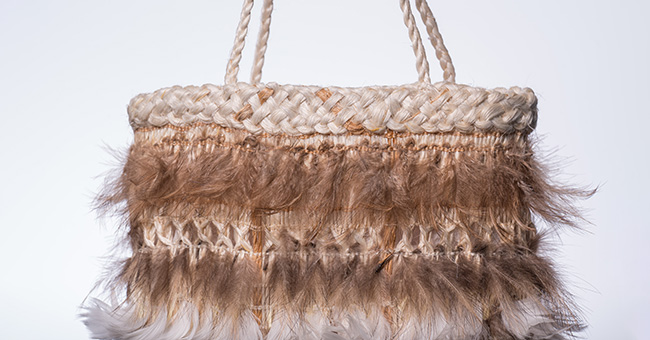 A woven kete decorated with feathers, photographed against a white background