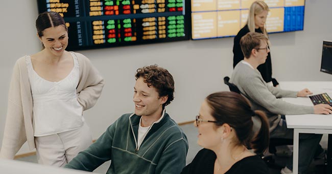 A group of students working at computers in the Bloomberg Markets Lab, Otago Business School, with data on large screens in the background