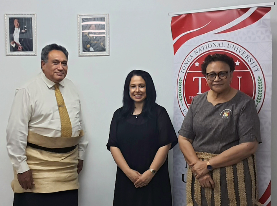 A group of people - two women and one man dressed in traditional Tongan attire, standing in front of a pull-up banner.