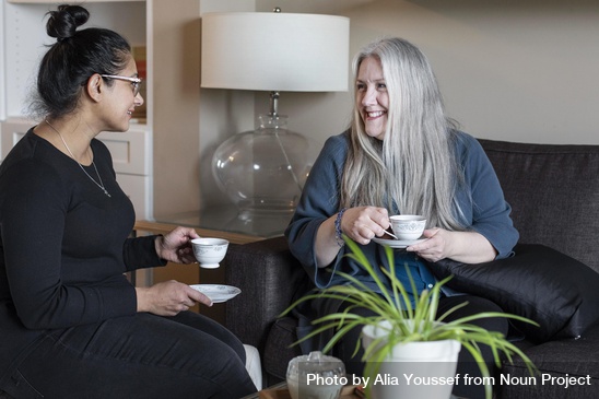 Two smiling women sit on a couch with cups of tea. Photo by Alia Youssef from Noun Project.