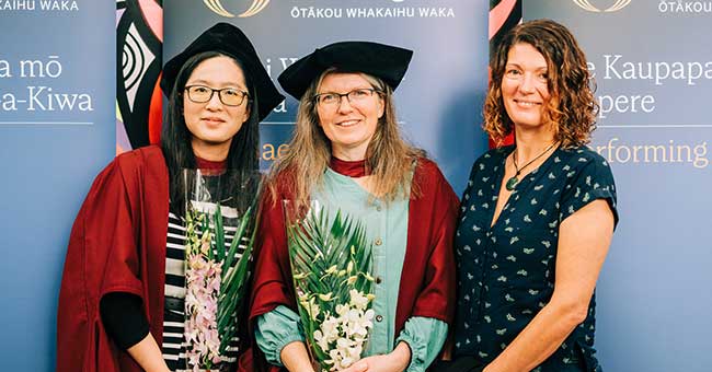 Three woman standing next to each other, two in graduation attire, in front of University of Otago banners