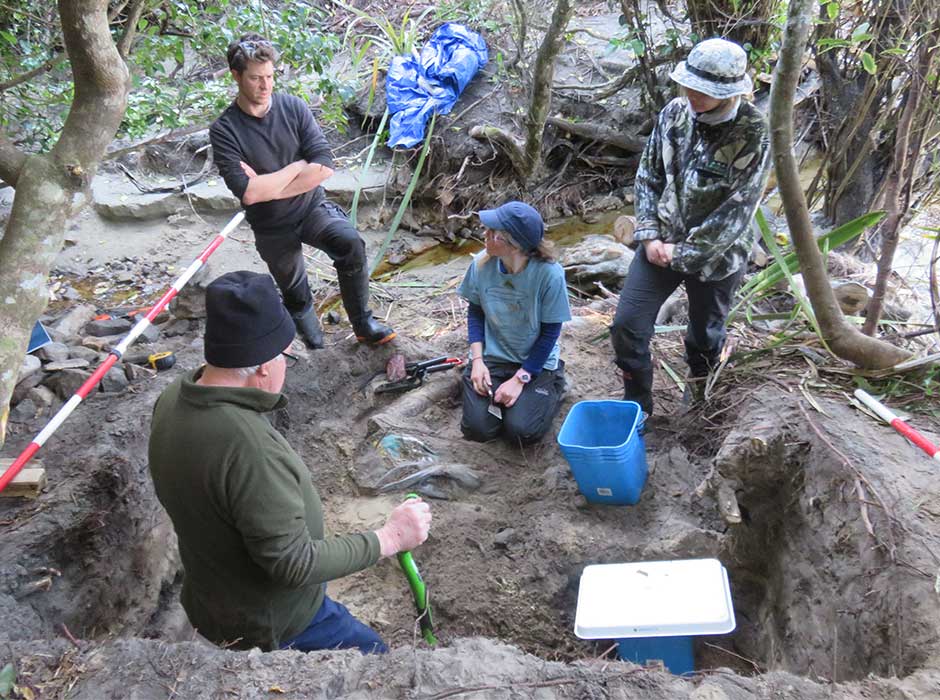 One person seated, with four others standing around them around an excavation site