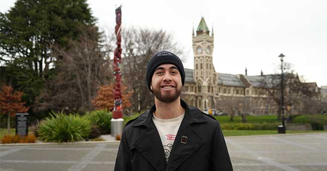 a young man standing in front of the Pou and Clocktower, smiling on an overcast day