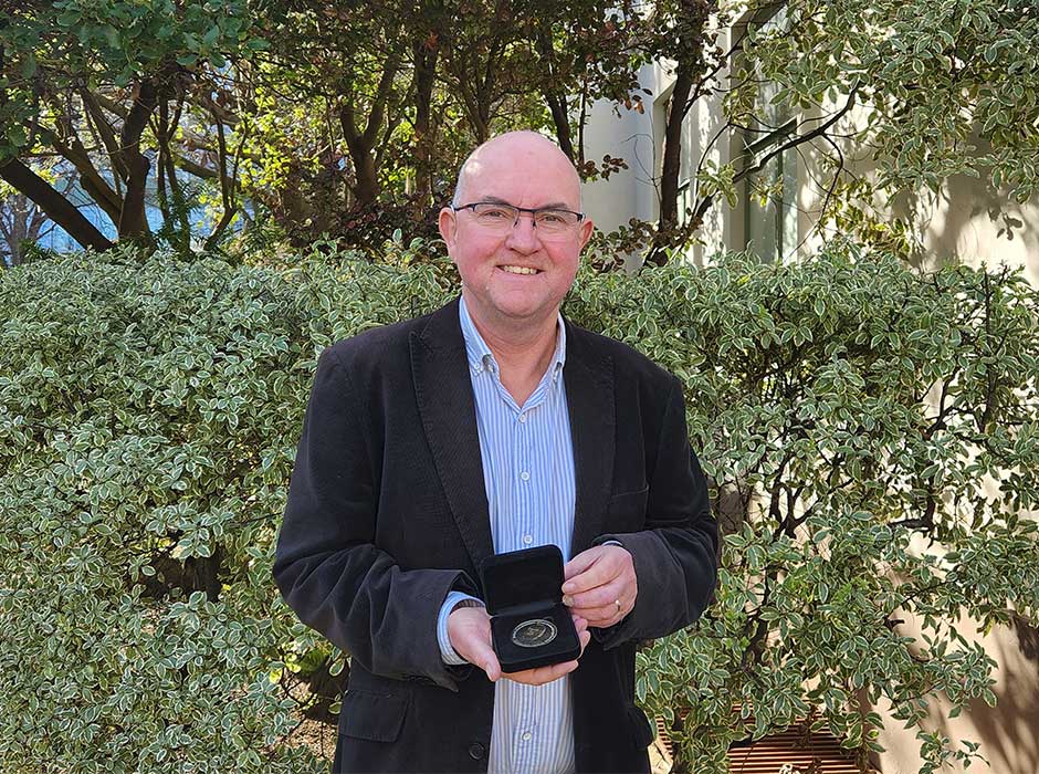 A man, from the waist up, standing in front of greenery smiling and holding a small box open with a medal inside it