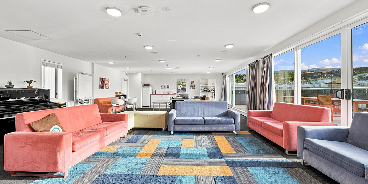 Shared lounge with sofas, tables, a piano and large windows with city views.
