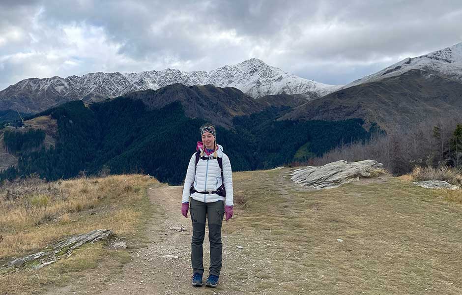 A woman standing on a tussocky area with snowy mountains in the background and a cloudy grey sky