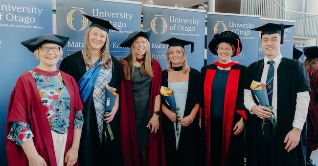 A group of six people dressed in black and scarlet academic regalia stand in front of blue University of Otago banners.