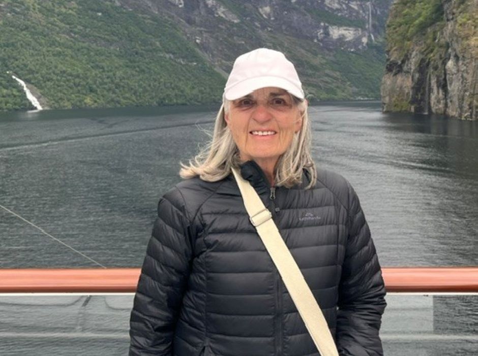 a woman standing on a boat in a black puffer jacket wearing a white baseball cap with an unspecified fjord behind her