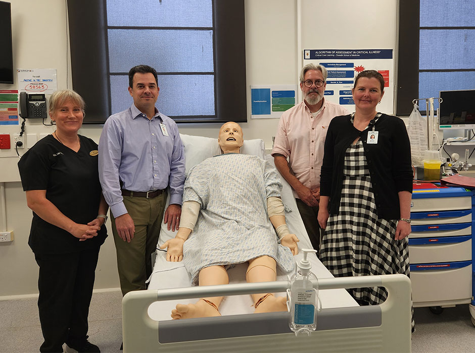 Four people standing either side of a hospital bed with a dummy in it