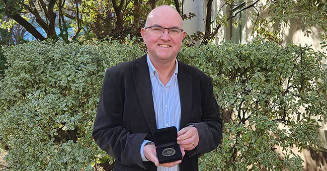 A man, from the waist up, standing in front of greenery smiling and holding a small box open with a medal inside it