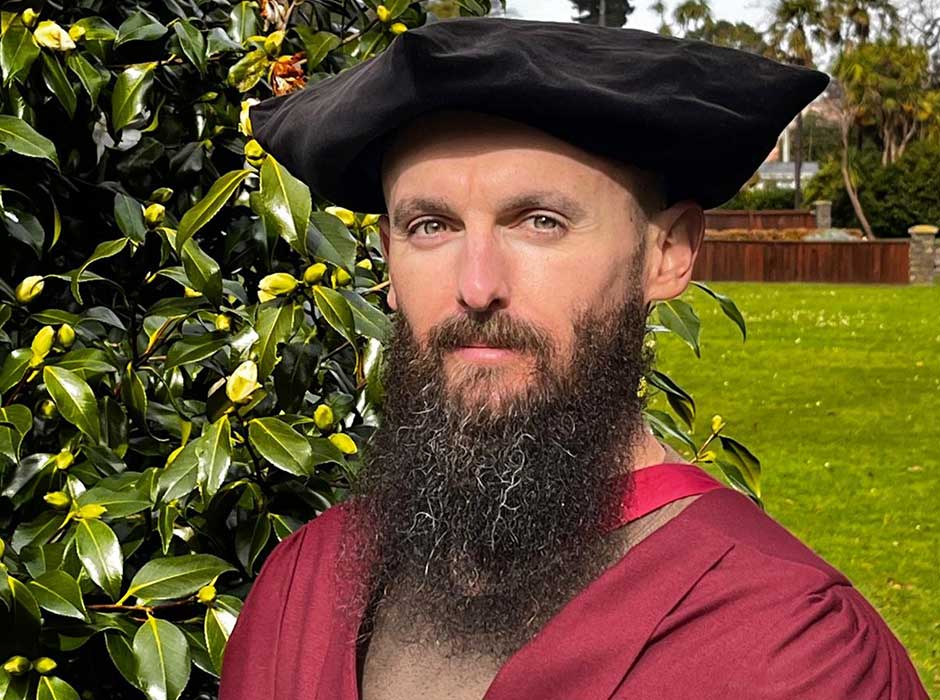 A man stands in the botanical gardens wearing a maroon graduation gown and black velvet graduation cap 