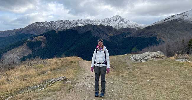 A woman standing on a tussocky area with snowy mountains in the background and a cloudy grey sky
