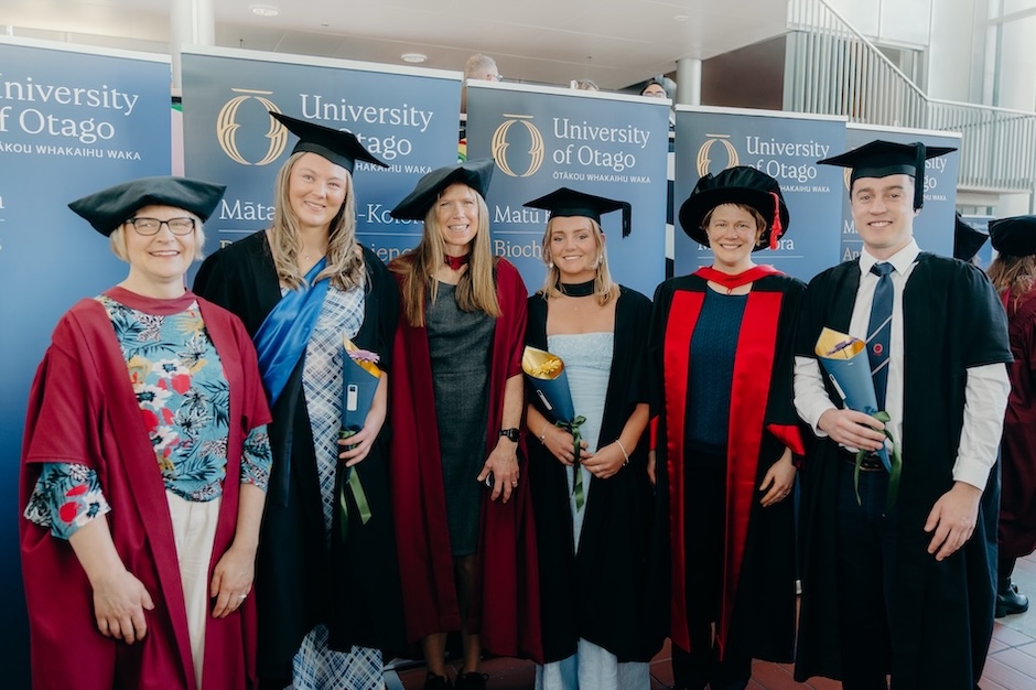 A group of six people dressed in black and scarlet academic regalia stand in front of blue University of Otago banners.