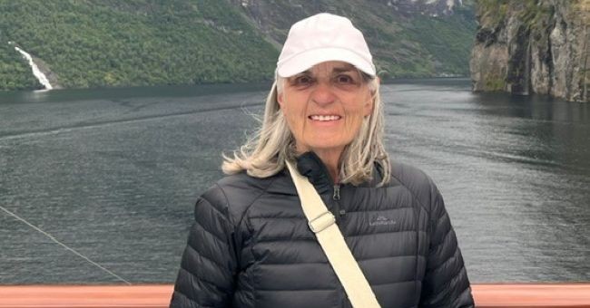 a woman standing on a boat in a black puffer jacket wearing a white baseball cap with an unspecified fjord behind her