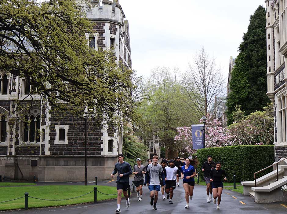 a group of people running towards the camera from a quad area surrounded by old stone buildings, leafy trees