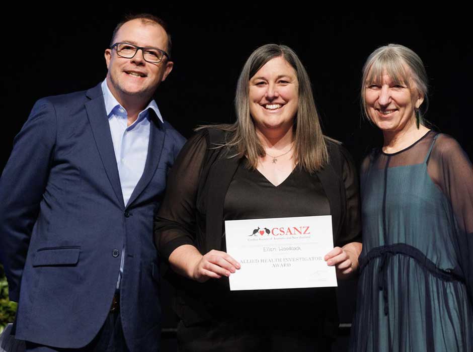 Three people standing smiling one in the centre holding an award 