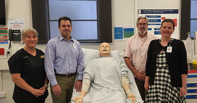 Four people standing either side of a hospital bed with a dummy in it