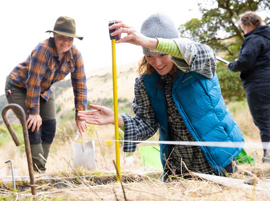 A woman standing in an excavation site, measuring, with two woman standing in the background 