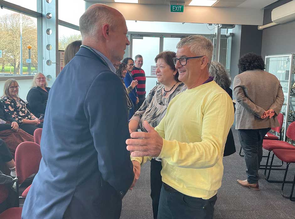 Two men greeting each other in a room with other people milling around behind thm