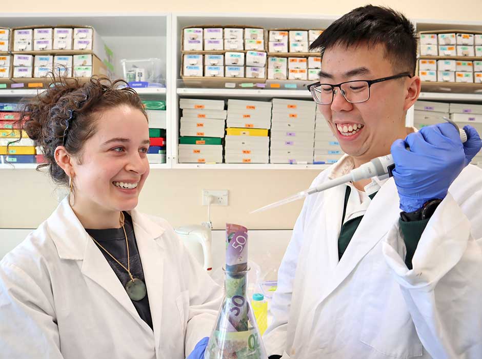 A young woman and a young man standing in a lab, wearing lab coats and holding up equipment