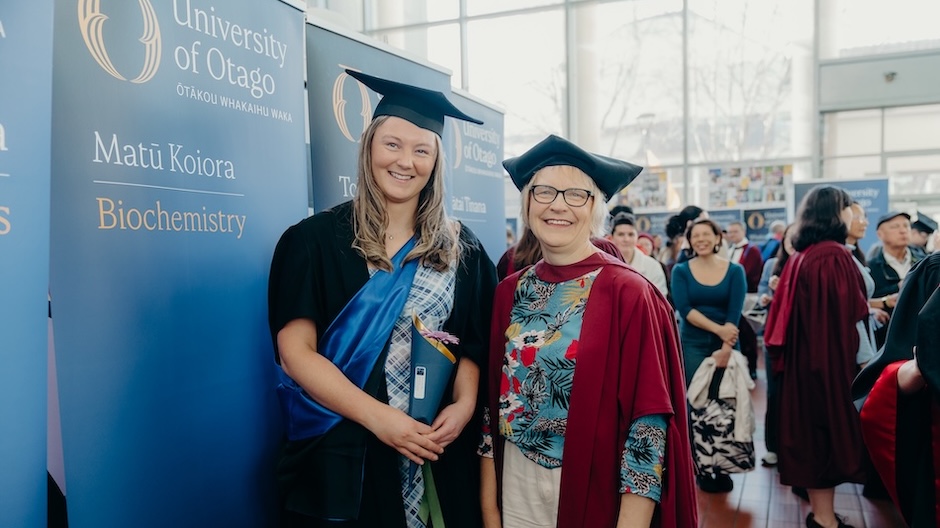 Two women dressed in black, blue and scarlet academic regalia stand in front of blue University of Otago banners and a crowd of people.