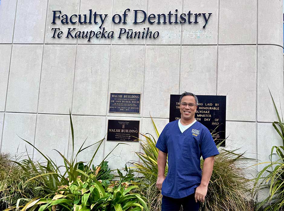 A man wearing blue scrubs standing in front of the faculty of dentistry sign