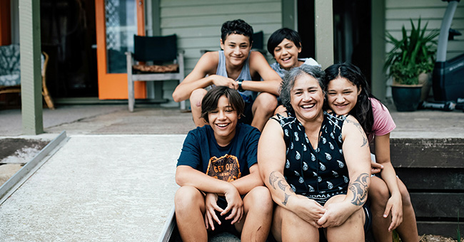 A wahine Māori and four tamariki, sitting together in front of a house and smiling