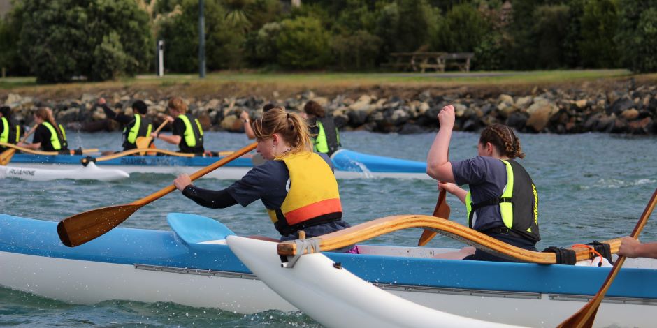 A close-up of two students paddling a blue waka, or outrigger canoe, with another waka full of students in the background.