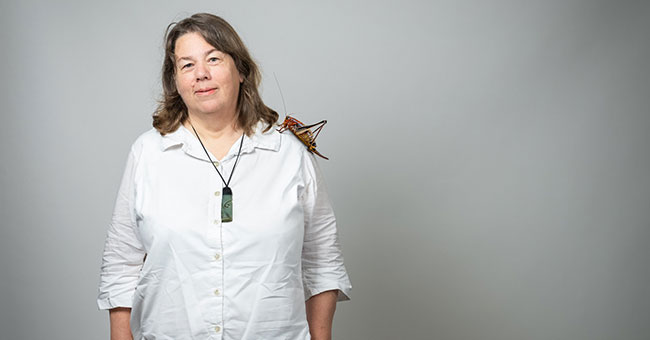 A woman wearing jeans, a white shirt and a greenstone necklace, standing in front of a blank backdrop
