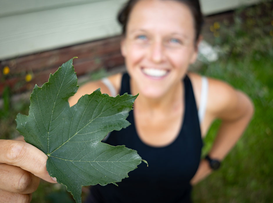 A blurry photo of a woman looking up at the camera smiling, holding a sycamore leaf in her hand that is in focus