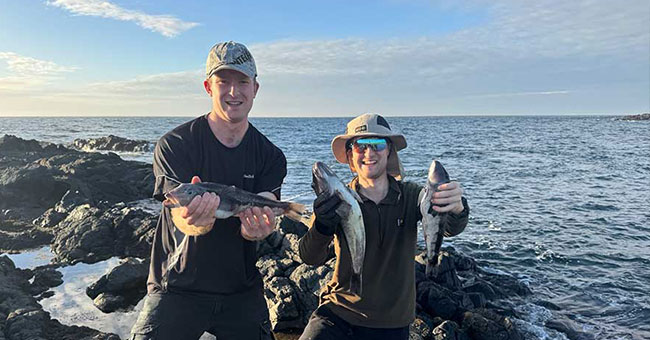 Two men, standing on a rocky outcrop holding fish, with the sea behind them.