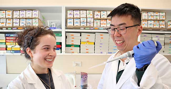 A young woman and a young man standing in a lab, wearing lab coats and holding up equipment