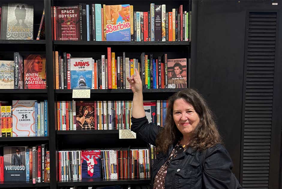 A women standing in front of a book case in a store, pointing to a book up on shelf above her