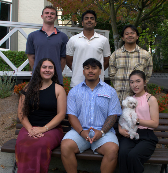 A group photograph of six smiling young people and a small white dog in an outdoor garden area.