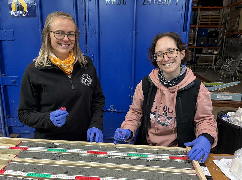 Two women standing behind a bench wearing gloves, with samples of mud on the table