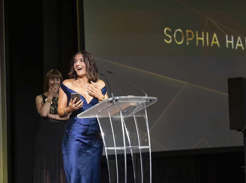 A woman in a blue dress standing at a podium with another woman in a black dress looks on.
