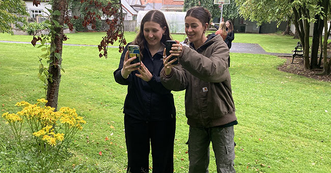 Two young women take photos of plant life with a mobile phone.