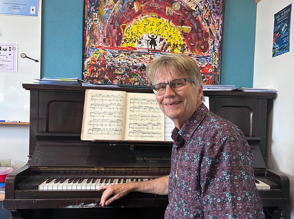A man sat at a piano, with a painting of an orchestra behind him