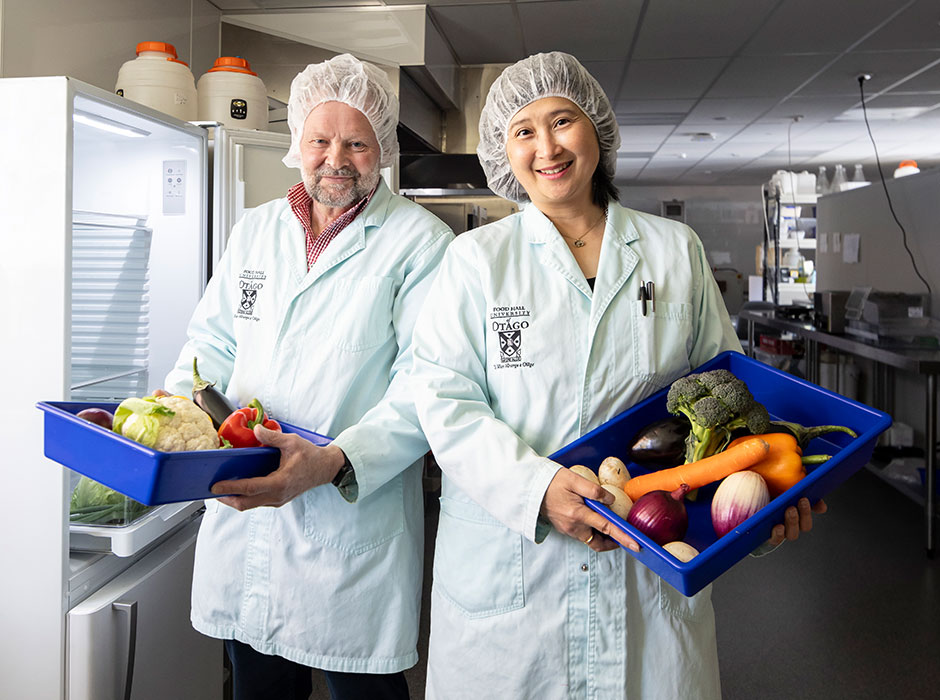 two people standing in lab coats, next to an open fridge and holding containers with vegetables in them