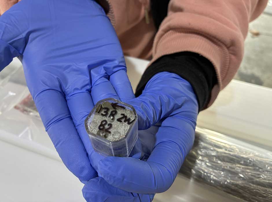 A pair of gloved hands holding a plastic container about an 1 x 1 x 1 inch in size, containing  sample of mud 