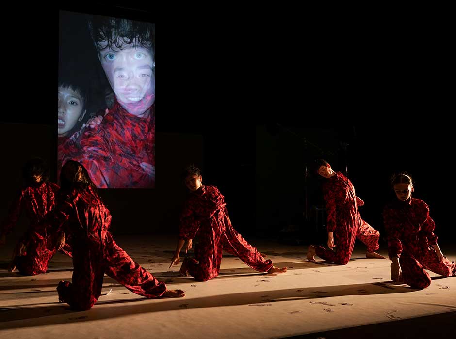 Six people in red costumes dancing on stage on their knees with a image of two people projected behind them