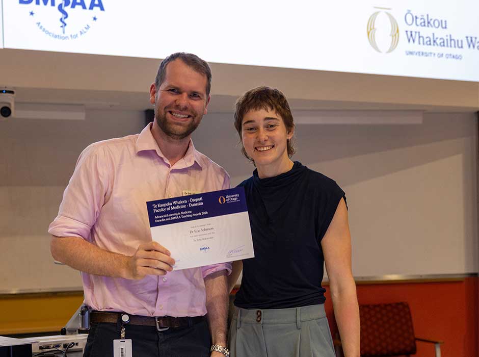 A man stands, smiling, holding a certificate with a woman standing next to him, also smiling