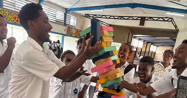 A group of young adults standing around a table, laughing and playing jenga 