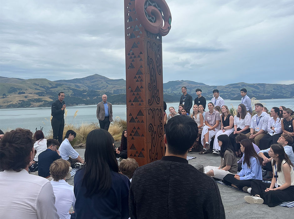 a man speaks, surrounded by young people, on the edge of Akaroa Harbour